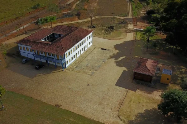 Imagem aérea da Fazenda da Vargem, em Nova Era, destacando um casarão colonial de dois pavimentos com telhado de barro, localizado em uma ampla área rural. Ao redor da construção há pátios, árvores e estradas internas, evidenciando o valor histórico e cultural do local.