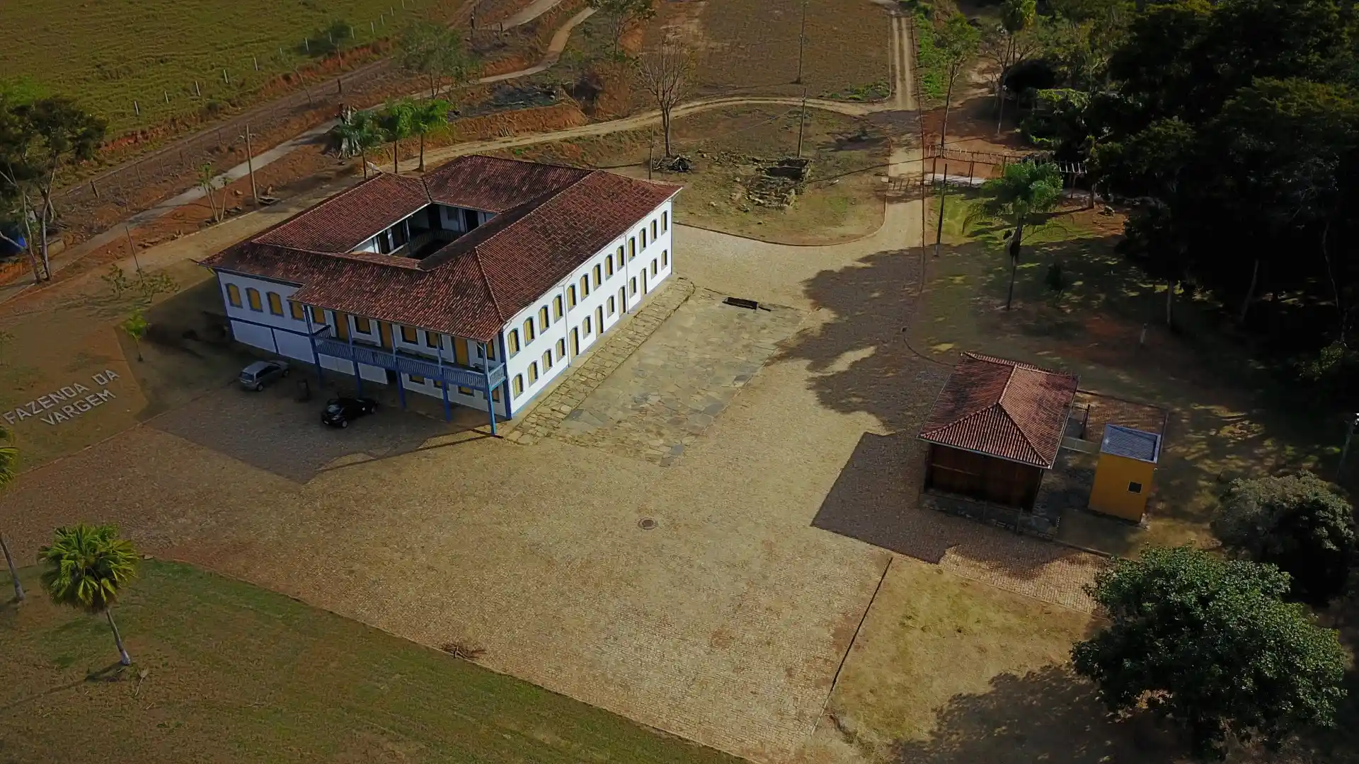 Imagem aérea da Fazenda da Vargem, em Nova Era, destacando um casarão colonial de dois pavimentos com telhado de barro, localizado em uma ampla área rural. Ao redor da construção há pátios, árvores e estradas internas, evidenciando o valor histórico e cultural do local.