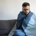 Homem sentado em um sofá, enrolado em um cobertor, demonstrando sensação de frio. A imagem ilustra o clima de baixas temperaturas previsto para a região, conforme a previsão do Inmet para João Monlevade e cidades do Médio Piracicaba.