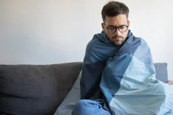 Homem sentado em um sofá, enrolado em um cobertor, demonstrando sensação de frio. A imagem ilustra o clima de baixas temperaturas previsto para a região, conforme a previsão do Inmet para João Monlevade e cidades do Médio Piracicaba.