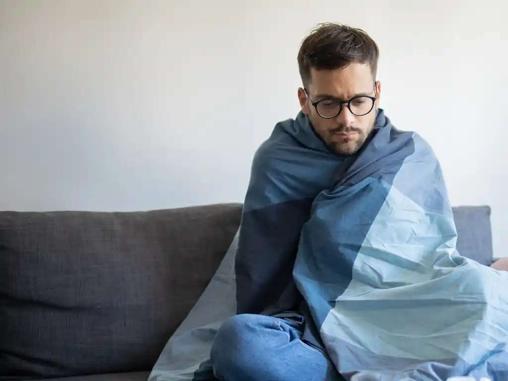 Homem sentado em um sofá, enrolado em um cobertor, demonstrando sensação de frio. A imagem ilustra o clima de baixas temperaturas previsto para a região, conforme a previsão do Inmet para João Monlevade e cidades do Médio Piracicaba.