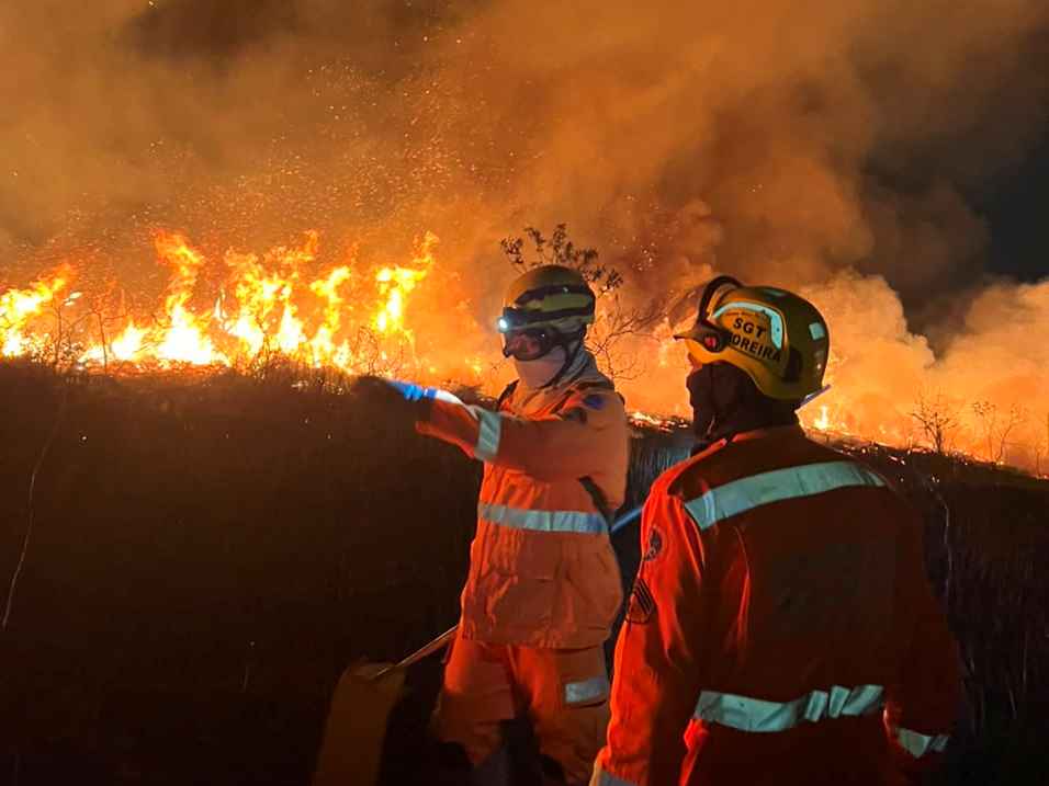 Dois integrantes do Corpo de Bombeiros observam e combatem um incêndio florestal de grandes proporções durante a noite. Ao fundo, uma extensa linha de chamas consome a vegetação, produzindo intensa fumaça e iluminando a cena com tons alaranjados.