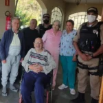 Policiais militares, profissionais de uma instituição e idosos posam para foto durante visita da Operação Virtude 2026 em uma das instituições de acolhimento para idosos de João Monlevade e região. Um idoso está sentado em cadeira de rodas, enquanto os demais participantes permanecem ao seu redor em área coberta da unidade.