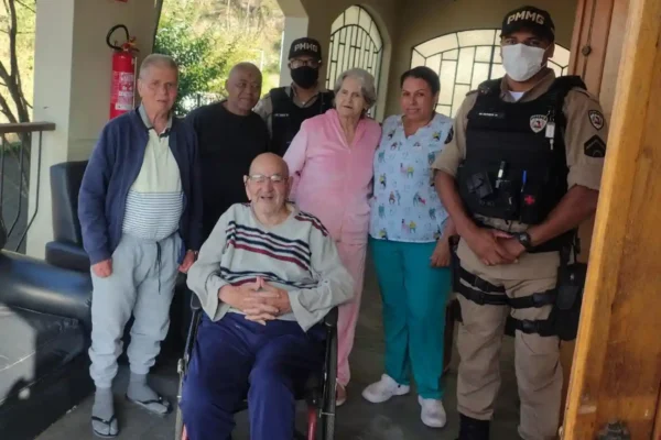 Policiais militares, profissionais de uma instituição e idosos posam para foto durante visita da Operação Virtude 2026 em uma das instituições de acolhimento para idosos de João Monlevade e região. Um idoso está sentado em cadeira de rodas, enquanto os demais participantes permanecem ao seu redor em área coberta da unidade.
