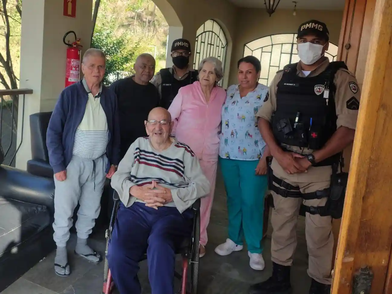 Policiais militares, profissionais de uma instituição e idosos posam para foto durante visita da Operação Virtude 2026 em uma das instituições de acolhimento para idosos de João Monlevade e região. Um idoso está sentado em cadeira de rodas, enquanto os demais participantes permanecem ao seu redor em área coberta da unidade.