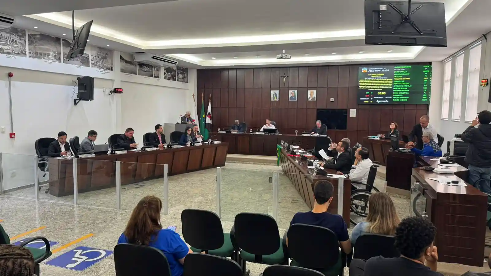 Imagem mostra os vereadores reunidos no plenário da Câmara Municipal durante sessão legislativa. Os parlamentares estão sentados em seus respectivos lugares enquanto acompanham a votação de projetos de lei. Ao fundo, um painel eletrônico exibe informações da reunião, e parte do público acompanha os debates nas cadeiras destinadas aos visitantes.