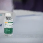 Frasco da vacina contra a dengue do Instituto Butantan em destaque sobre uma mesa. O imunizante teve a aplicação suspensa temporariamente pelo Ministério da Saúde após a investigação de casos de reações graves e duas mortes suspeitas.
