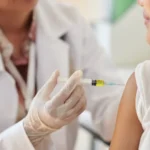 Close-up de um profissional de saúde usando luvas e aplicando uma vacina no braço de uma criança. A cena representa a vacinação de crianças, fundamental para a proteção contra doenças imunopreveníveis e para a atualização da caderneta vacinal.