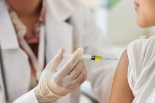 Close-up de um profissional de saúde usando luvas e aplicando uma vacina no braço de uma criança. A cena representa a vacinação de crianças, fundamental para a proteção contra doenças imunopreveníveis e para a atualização da caderneta vacinal.