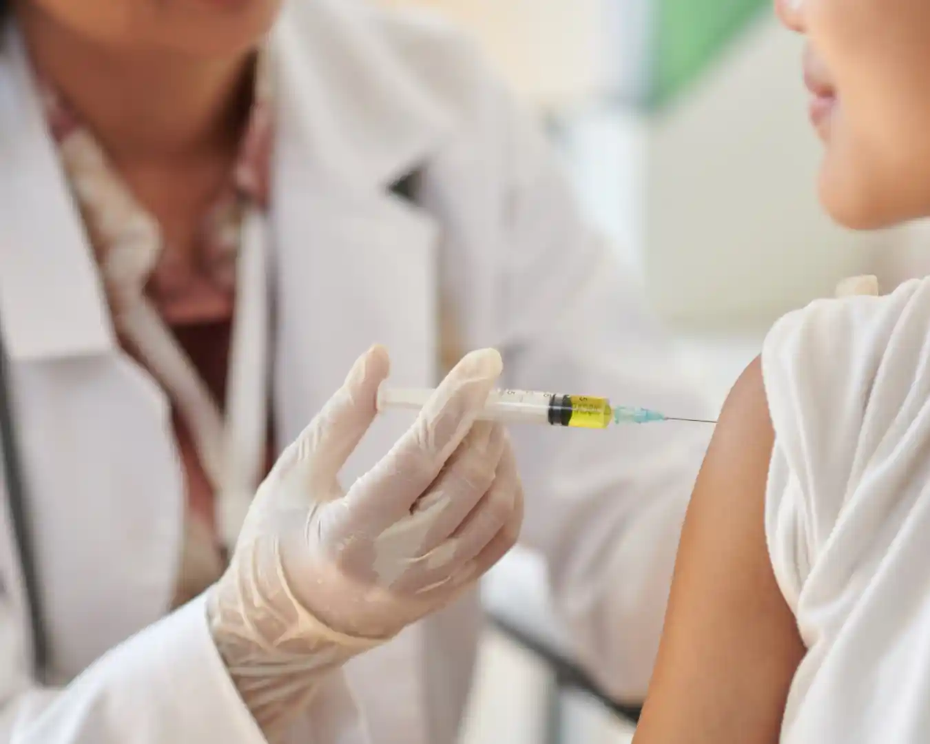 Close-up de um profissional de saúde usando luvas e aplicando uma vacina no braço de uma criança. A cena representa a vacinação de crianças, fundamental para a proteção contra doenças imunopreveníveis e para a atualização da caderneta vacinal.