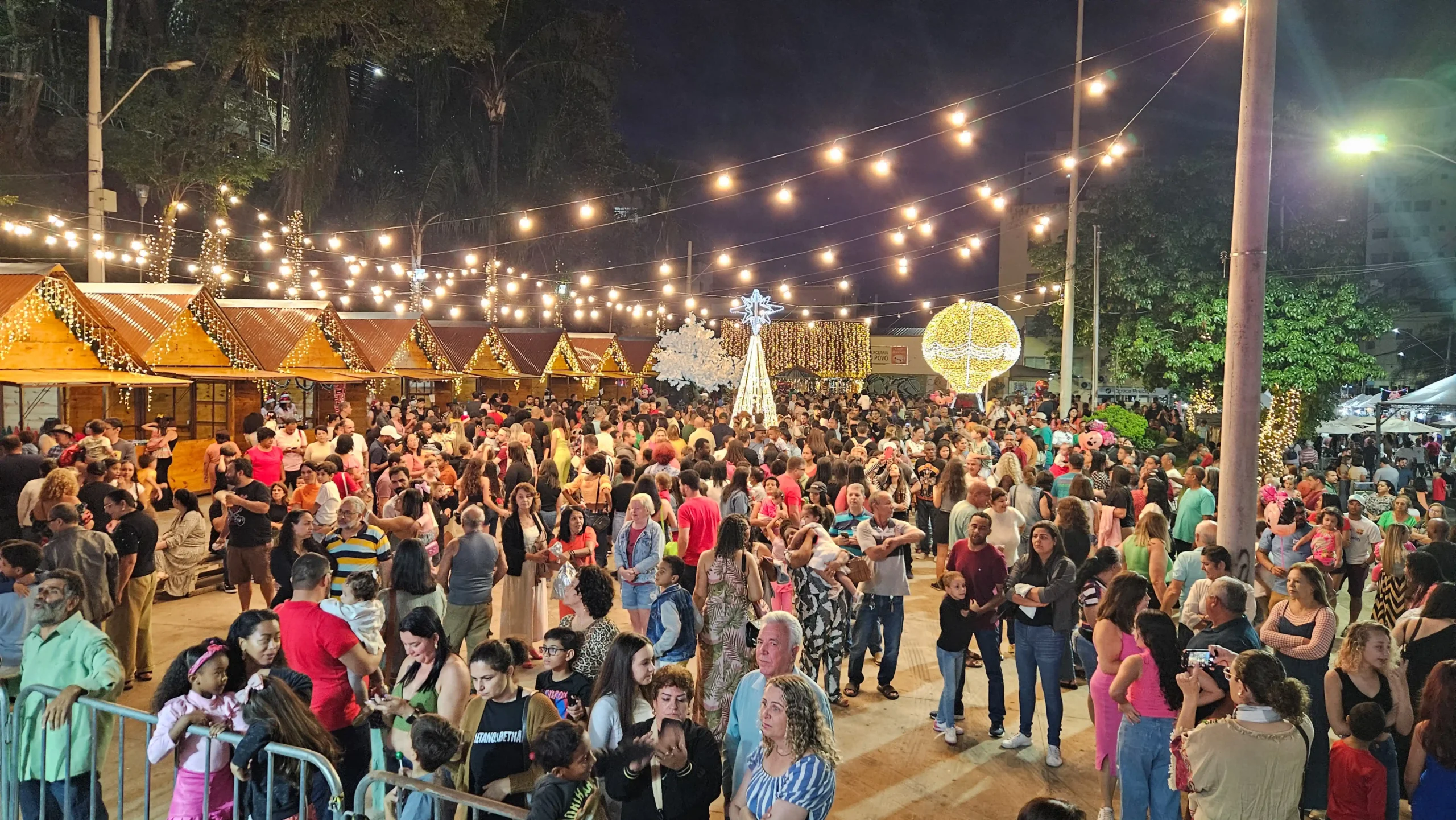Grande público visita o Mercado de Natal organizado pela Casa de Cultura em João Monlevade, com chalés iluminados, decoração natalina, luzes suspensas e famílias circulando pelo evento durante a noite.