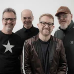 Quatro integrantes da banda Nenhum de Nós sorriem para a câmera diante de um fundo branco. A imagem divulga a participação do grupo no Festival Gastronômico de Rio Piracicaba. Um dos músicos usa camiseta preta com estrela branca, outro veste blazer escuro, enquanto os demais aparecem com roupas escuras, óculos e boné.