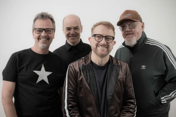 Quatro integrantes da banda Nenhum de Nós sorriem para a câmera diante de um fundo branco. A imagem divulga a participação do grupo no Festival Gastronômico de Rio Piracicaba. Um dos músicos usa camiseta preta com estrela branca, outro veste blazer escuro, enquanto os demais aparecem com roupas escuras, óculos e boné.