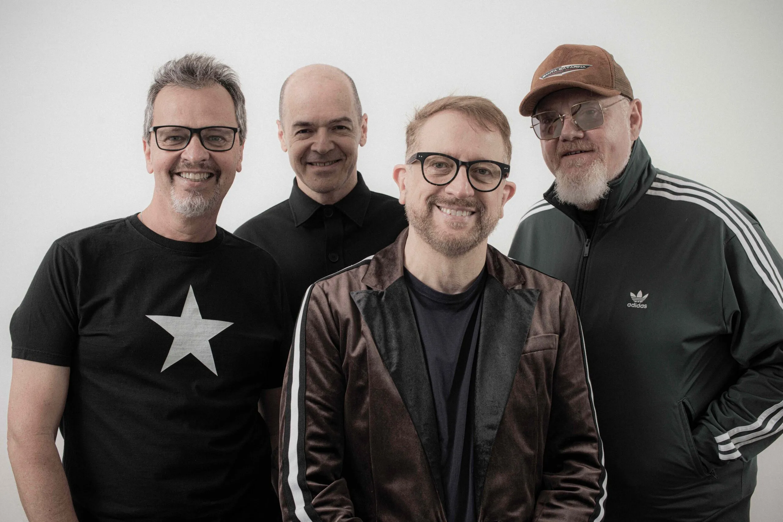 Quatro integrantes da banda Nenhum de Nós sorriem para a câmera diante de um fundo branco. A imagem divulga a participação do grupo no Festival Gastronômico de Rio Piracicaba. Um dos músicos usa camiseta preta com estrela branca, outro veste blazer escuro, enquanto os demais aparecem com roupas escuras, óculos e boné.