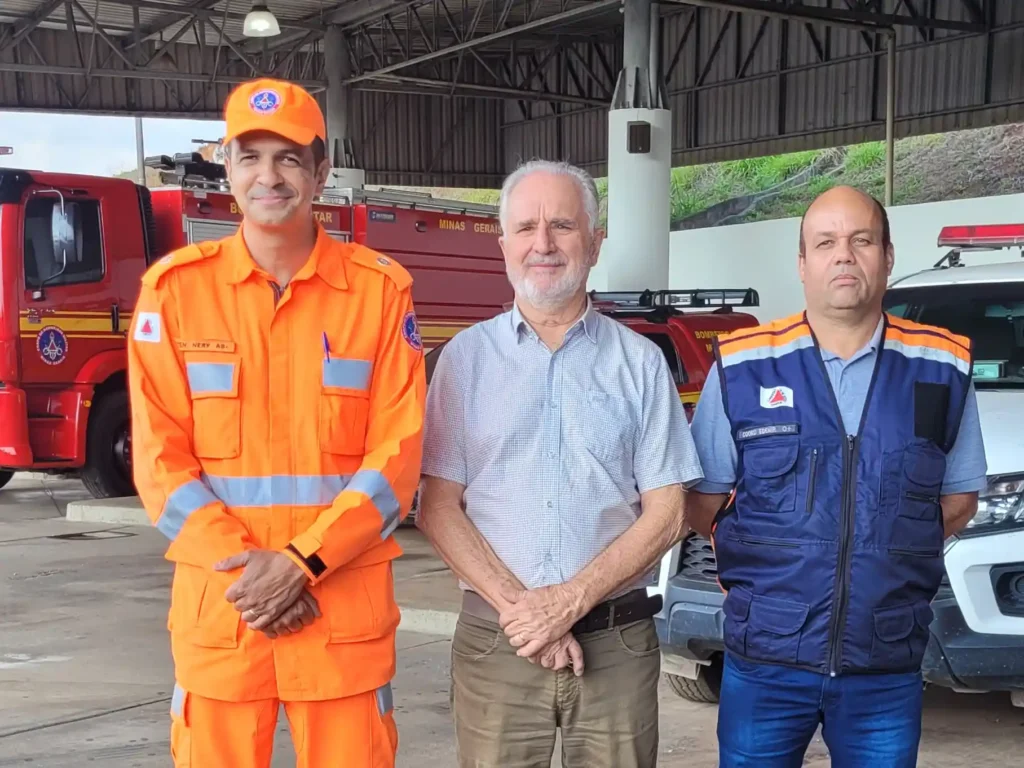 Três representantes posam para foto nas novas instalações do Centro de Urgência de João Monlevade. À esquerda, um militar do Corpo de Bombeiros Militar de Minas Gerais veste uniforme laranja. Ao centro, está o prefeito Dr. Laércio Ribeiro. À direita, o coordenador da Defesa Civil Municipal utiliza colete azul. Ao fundo, aparecem viaturas do Corpo de Bombeiros e da Defesa Civil estacionadas no espaço que passa a abrigar os serviços de emergência no município.