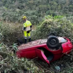 Integrantes do SEVOR e do Corpo de Bombeiros observam um carro vermelho capotado em uma ribanceira coberta por vegetação, durante operação de busca em João Monlevade. Nenhuma vítima foi encontrada no local.