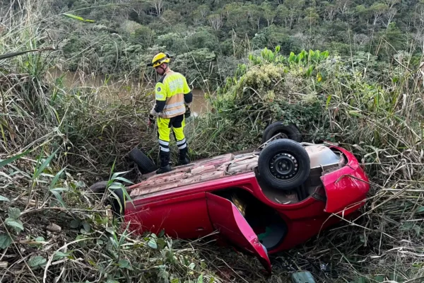 Integrantes do SEVOR e do Corpo de Bombeiros observam um carro vermelho capotado em uma ribanceira coberta por vegetação, durante operação de busca em João Monlevade. Nenhuma vítima foi encontrada no local.