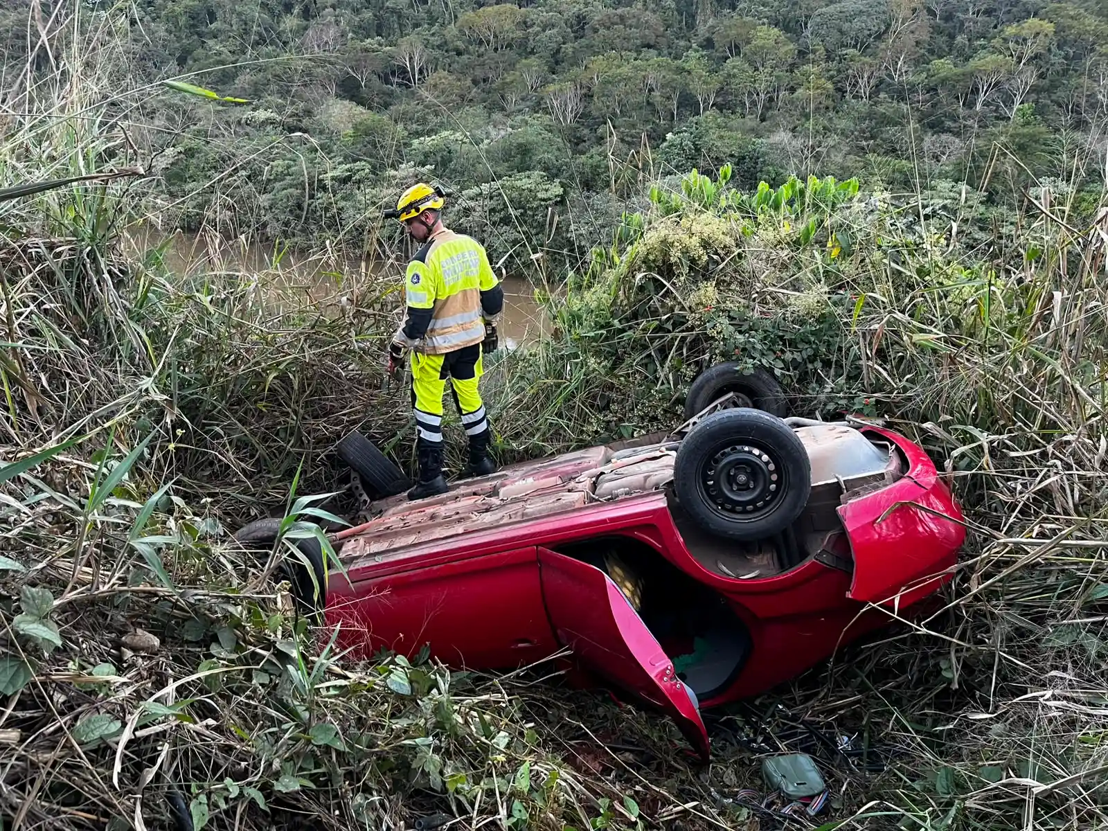 Integrantes do SEVOR e do Corpo de Bombeiros observam um carro vermelho capotado em uma ribanceira coberta por vegetação, durante operação de busca em João Monlevade. Nenhuma vítima foi encontrada no local.