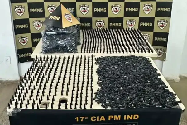 Fotografia da Polícia Militar em João Monlevade exibe mesa com 1.629 pinos de cocaína apreendidos, pacotes de pinos vazios e materiais relacionados ao tráfico de drogas. Ao fundo, há um painel com os brasões da PMMG utilizado para apresentação oficial da ocorrência.