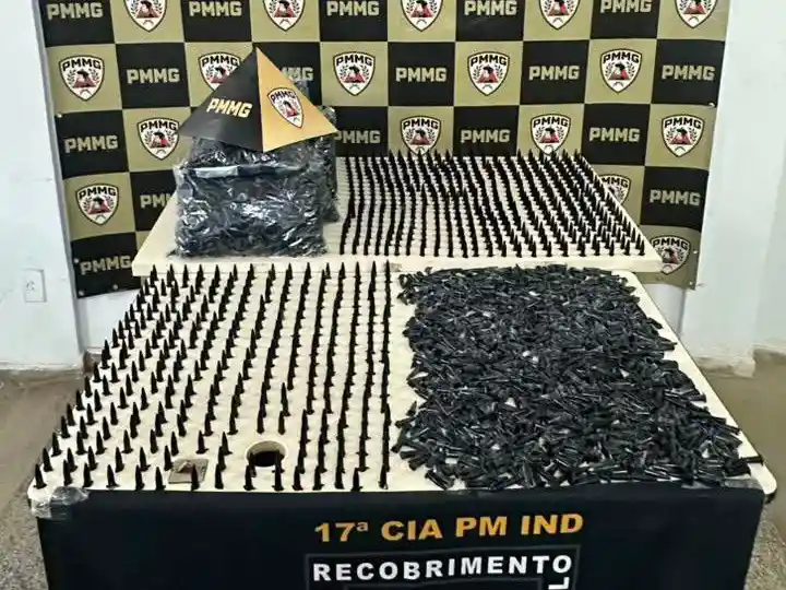 Fotografia da Polícia Militar em João Monlevade exibe mesa com 1.629 pinos de cocaína apreendidos, pacotes de pinos vazios e materiais relacionados ao tráfico de drogas. Ao fundo, há um painel com os brasões da PMMG utilizado para apresentação oficial da ocorrência.