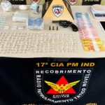 Mesa de exposição do Tático Móvel da Polícia Militar com drogas apreendidas, revólver calibre .38, munições, balança de precisão, celulares e dinheiro durante operação em João Monlevade.