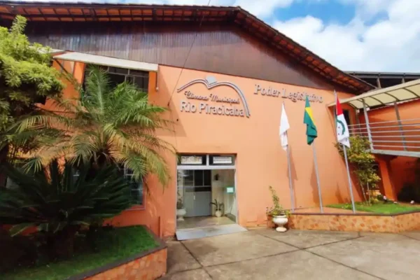 Fachada da Câmara de Rio Piracicaba, prédio de cor laranja com bandeiras em frente. A imagem ilustra notícia sobre o concurso da Câmara de Rio Piracicaba, que oferece cinco vagas efetivas para diferentes níveis de escolaridade.