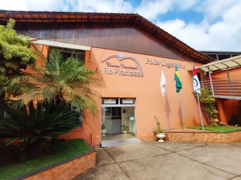 Fachada da Câmara de Rio Piracicaba, prédio de cor laranja com bandeiras em frente. A imagem ilustra notícia sobre o concurso da Câmara de Rio Piracicaba, que oferece cinco vagas efetivas para diferentes níveis de escolaridade.