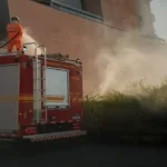 Viatura do Corpo de Bombeiros estacionada ao lado de uma área de vegetação em chamas no bairro Aclimação, em João Monlevade. Um bombeiro atua sobre o veículo utilizando mangueira para controlar o incêndio, enquanto uma densa fumaça se espalha próximo a um edifício residencial.