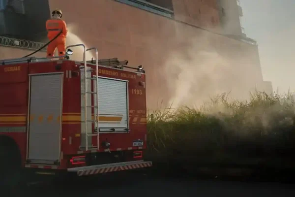 Viatura do Corpo de Bombeiros estacionada ao lado de uma área de vegetação em chamas no bairro Aclimação, em João Monlevade. Um bombeiro atua sobre o veículo utilizando mangueira para controlar o incêndio, enquanto uma densa fumaça se espalha próximo a um edifício residencial.