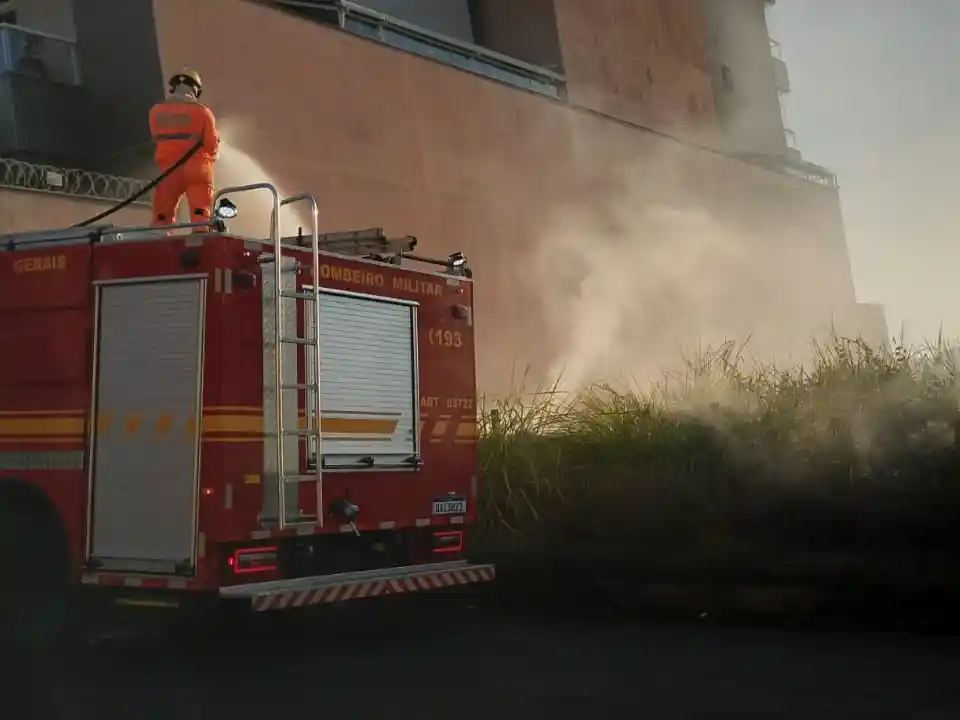 Viatura do Corpo de Bombeiros estacionada ao lado de uma área de vegetação em chamas no bairro Aclimação, em João Monlevade. Um bombeiro atua sobre o veículo utilizando mangueira para controlar o incêndio, enquanto uma densa fumaça se espalha próximo a um edifício residencial.