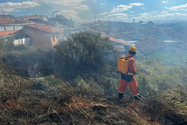 Bombeiro combate incêndio em vegetação no bairro Planalto, em João Monlevade. Ao fundo, casas da região aparecem envoltas por fumaça enquanto equipes trabalham para controlar as chamas próximas às residências.