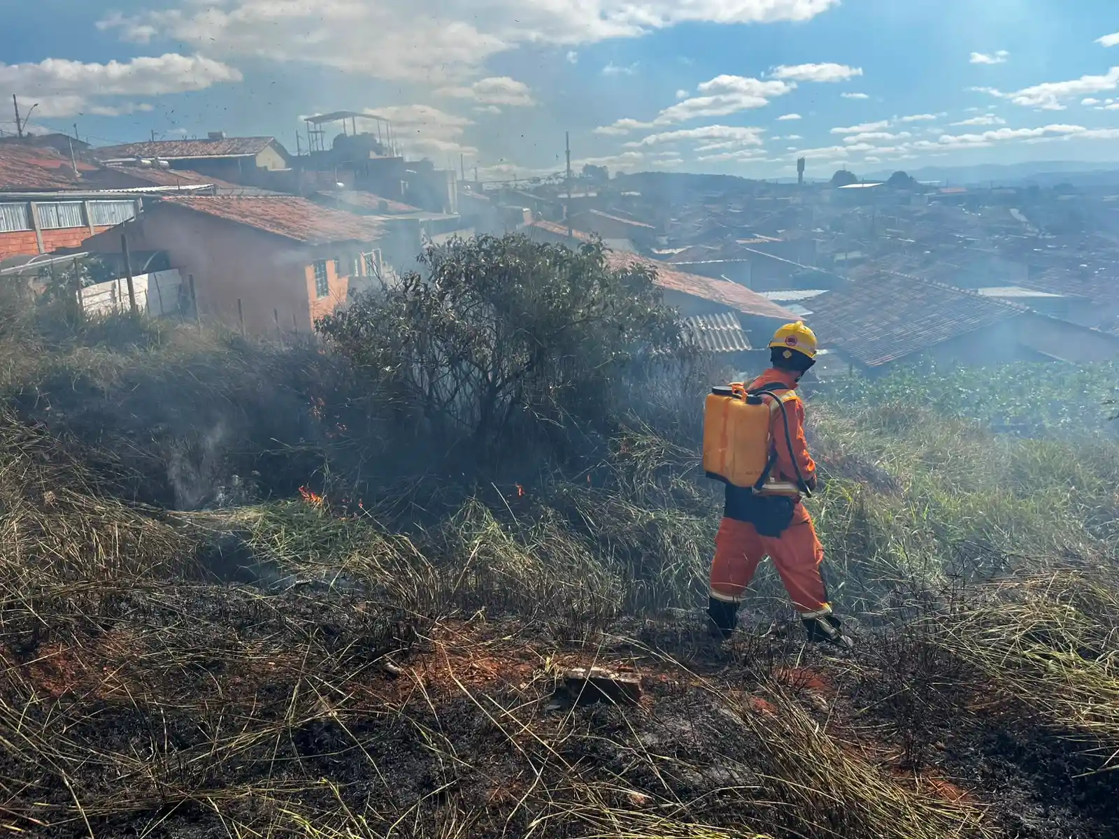 Bombeiro combate incêndio em vegetação no bairro Planalto, em João Monlevade. Ao fundo, casas da região aparecem envoltas por fumaça enquanto equipes trabalham para controlar as chamas próximas às residências.