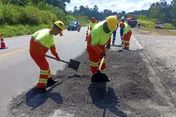 Trabalhadores uniformizados realizam reparos no asfalto da BR-381, utilizando pás para espalhar massa asfáltica em uma das faixas da rodovia. O local está sinalizado com cones, enquanto veículos passam pelo trecho em obras, que integra ações para melhorar o trânsito em João Monlevade.