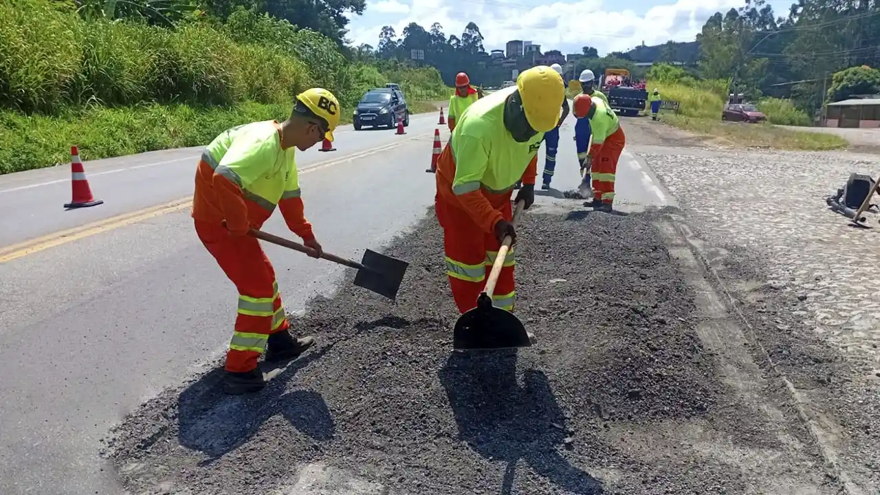 Trabalhadores uniformizados realizam reparos no asfalto da BR-381, utilizando pás para espalhar massa asfáltica em uma das faixas da rodovia. O local está sinalizado com cones, enquanto veículos passam pelo trecho em obras, que integra ações para melhorar o trânsito em João Monlevade.