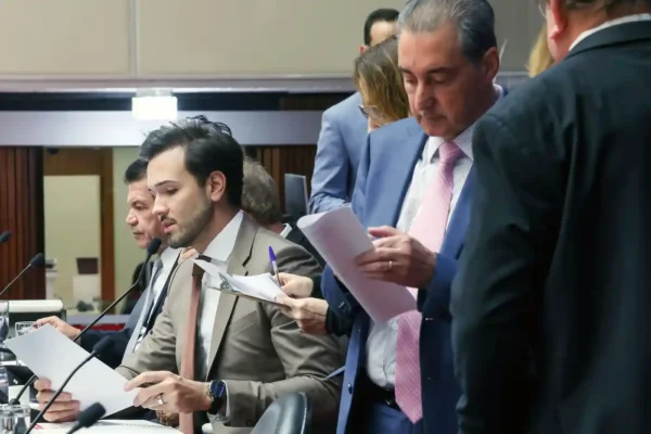 Parlamentares reunidos durante sessão da Assembleia Legislativa de Minas Gerais, com deputados analisando documentos no plenário. A imagem ilustra a votação do projeto que fixa teto para cachês artísticos e que será encaminhado ao governador de Minas Gerais para sanção.