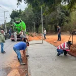 Trabalhadores realizam a concretagem de uma calçada às margens da Avenida Santa Cruz, em João Monlevade, durante a conclusão das obras de infraestrutura e drenagem no bairro Santa Cruz. Ao fundo, um caminhão-betoneira abastece o concreto enquanto equipes finalizam os serviços.