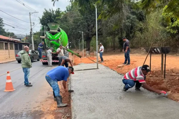 Trabalhadores realizam a concretagem de uma calçada às margens da Avenida Santa Cruz, em João Monlevade, durante a conclusão das obras de infraestrutura e drenagem no bairro Santa Cruz. Ao fundo, um caminhão-betoneira abastece o concreto enquanto equipes finalizam os serviços.