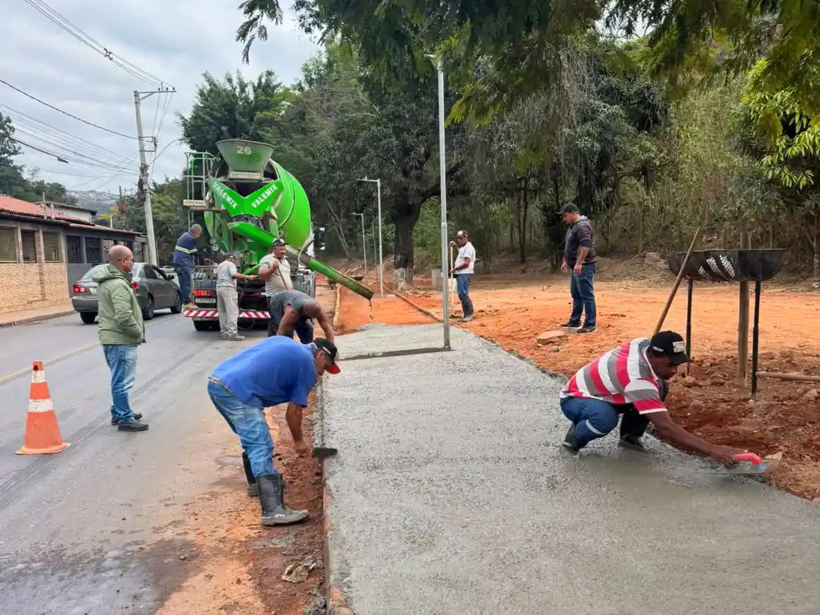 Trabalhadores realizam a concretagem de uma calçada às margens da Avenida Santa Cruz, em João Monlevade, durante a conclusão das obras de infraestrutura e drenagem no bairro Santa Cruz. Ao fundo, um caminhão-betoneira abastece o concreto enquanto equipes finalizam os serviços.