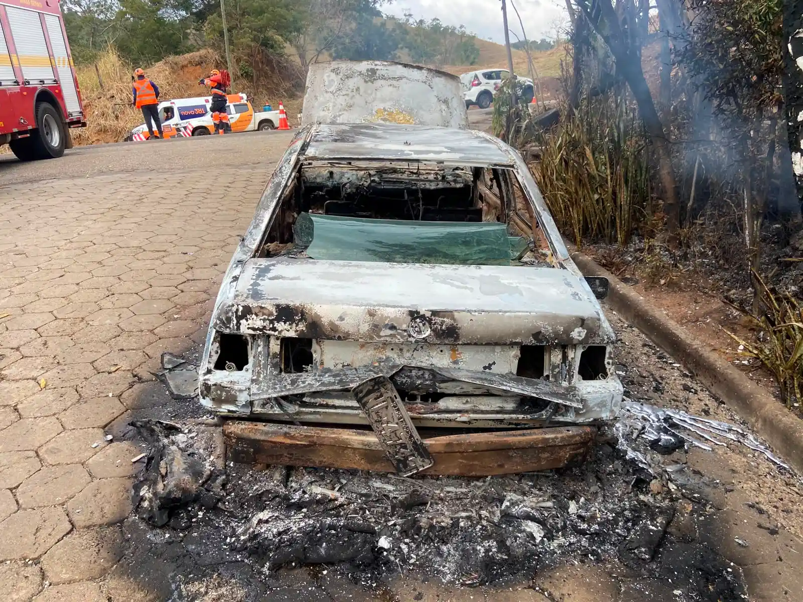 Carro Santana totalmente queimado após incêndio na BR-381, em Nova Era, com equipes do Corpo de Bombeiros e da Nova 381 atuando no local.
