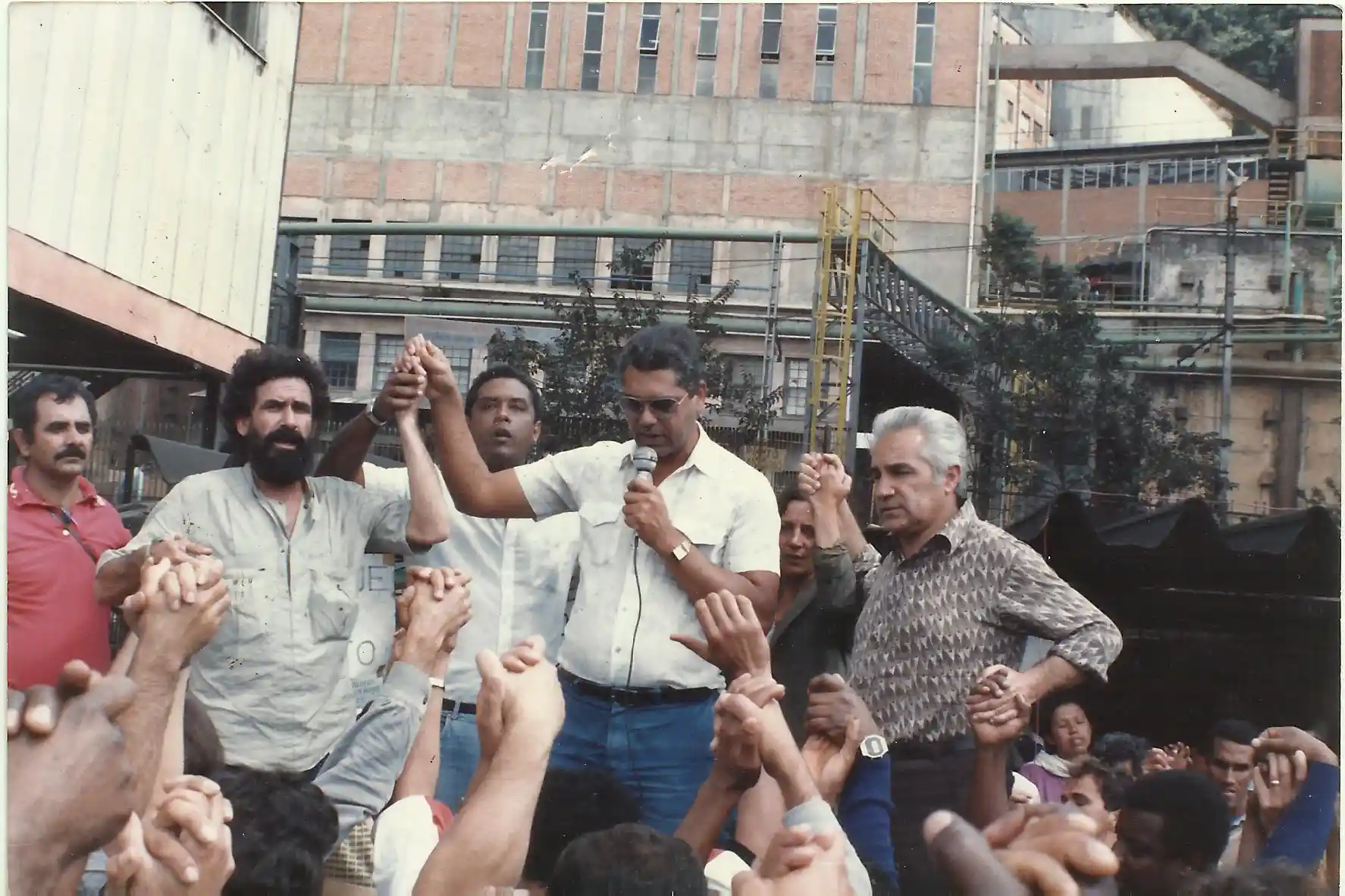 Greve de 1986 em João Monlevade com trabalhadores reunidos durante manifestação, levantando os braços em ato de mobilização e oração.